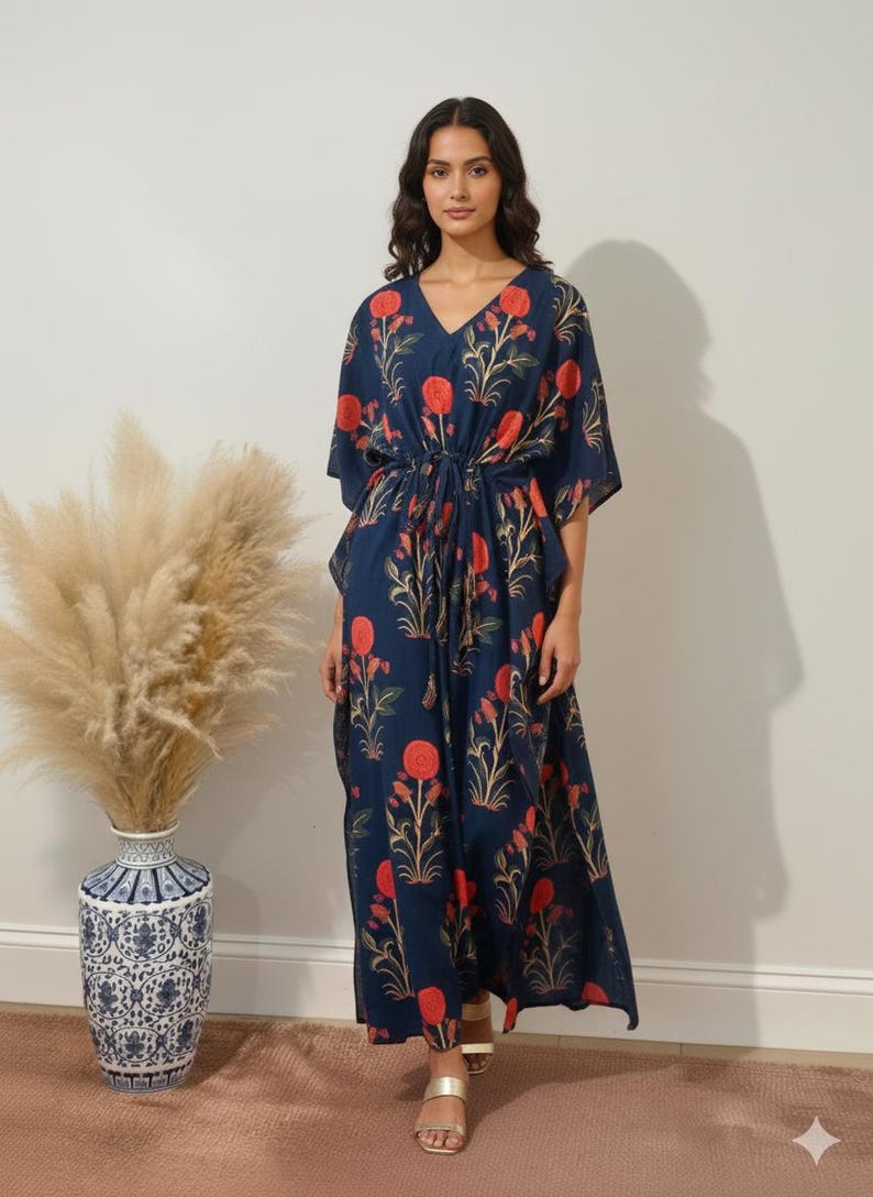 K&ouml;nnte beinhalten: Marineblaues Kaftankleid mit V-Ausschnitt und floralem Muster in Rot und Gold. Das Kleid hat eine Kordel in der Taille und kurze &Auml;rmel. Das Model tr&auml;gt goldene Sandalen.