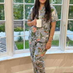 Op de afbeelding: Lichtblauwe pyjamaset met een botanische en dierenprint, inclusief giraffen en vogels. De set heeft een overhemd met witte bies en een bijpassende broek. Een witte mok met een berenontwerp wordt vastgehouden.