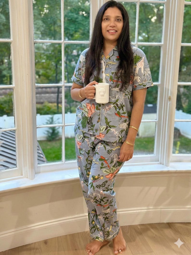 Op de afbeelding: Lichtblauwe pyjamaset met een botanische en dierenprint, inclusief giraffen en vogels. De set heeft een overhemd met witte bies en een bijpassende broek. Een witte mok met een berenontwerp wordt vastgehouden.