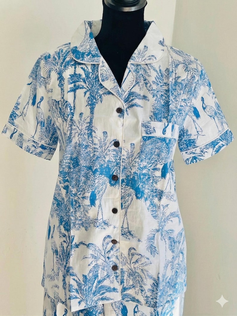 Op de afbeelding: Wit overhemd met een blauwe tropische print met palmbomen en vogels. Het shirt met korte mouwen heeft een reverskraag, een borstzak en donkerbruine knopen. Het shirt maakt deel uit van een pyjamaset.