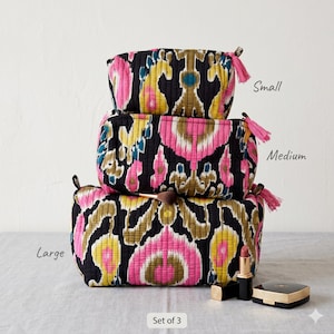 May include: Set of 3 quilted cosmetic bags in black, pink, yellow, and teal ikat pattern. The bags are labeled small, medium, and large. Each bag has a pink tassel and a zipper closure. A makeup brush, lipstick, and compact are next to the bags.