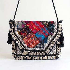 May include: A rectangular shoulder bag featuring a patchwork of colorful embroidered fabric panels in red, blue, and purple. The flap is decorated with rows of silver-toned metal coins and black fringe tassels on the sides, with a black braided strap.