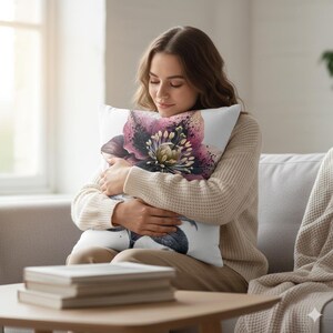 May include: A white decorative pillow featuring a large, detailed floral design in shades of purple, pink, and green. The pillow is rectangular and appears soft, with a close-up view of the flower's intricate details.