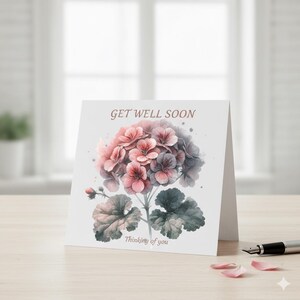 May include: A folded greeting card with a watercolor illustration of pink and purple flowers and green leaves. The text "GET WELL SOON" is at the top, and "Thinking of you" is at the bottom. A pen and petals are in the foreground.