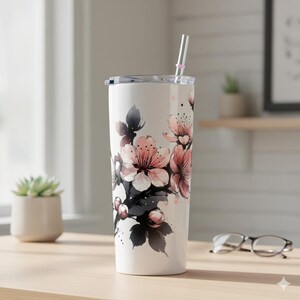 May include: A white tumbler with a floral design featuring pink and white blossoms and dark gray leaves. The tumbler has a clear lid and a clear straw with a pink ring. The tumbler is on a wooden surface.