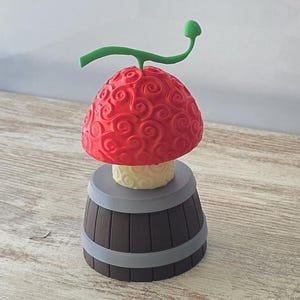 Puede incluir: Una figura con forma de fruta roja texturizada con un tallo verde y una base de color claro, colocada sobre una base en forma de barril marrón con bandas grises. La figura es una representación detallada y estilizada de una fruta ficticia.