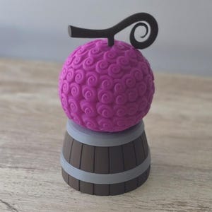 Puede incluir: Una fruta esférica de color magenta con un patrón arremolinado, coronada por un tallo negro y un remolino decorativo. Descansa sobre una base en forma de barril marrón con bandas grises, creando un diseño único y llamativo.