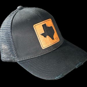 Op de afbeelding: Zwarte truckerpet met een versleten rand en een mesh achterkant. De voorkant heeft een vierkante, bruine leren patch met een zwarte omtrek van de staat Texas en de letters "TX".