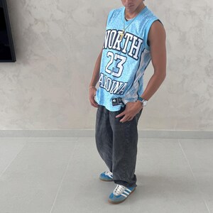 May include: A light blue basketball jersey with the text "NORTH 23 ANNA" in white and black. The jersey is paired with dark gray jeans and blue sneakers with white stripes.