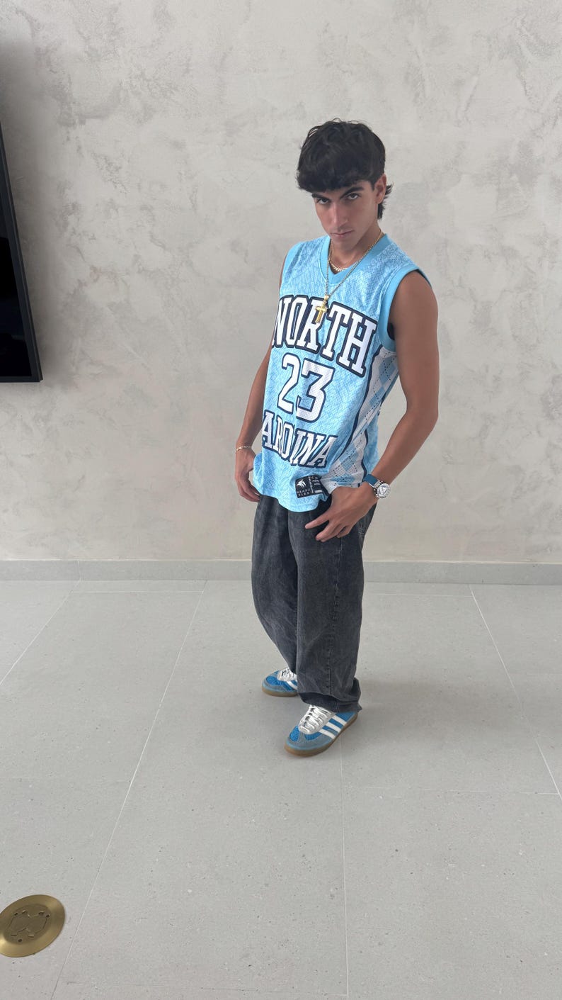 May include: A light blue basketball jersey with the text "NORTH 23 ANNA" in white and black. The jersey is paired with dark gray jeans and blue sneakers with white stripes.