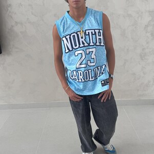 May include: A light blue basketball jersey with the text "NORTH 23 CAROLINA" is worn with dark gray jeans and blue sneakers. The jersey has a white and blue pattern. A gold necklace and watch are also visible.