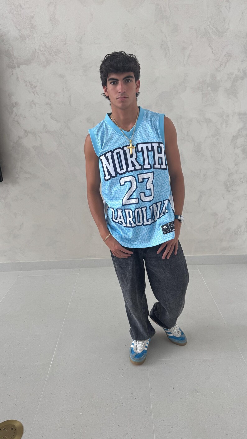May include: A light blue basketball jersey with the text "NORTH 23 CAROLINA" is worn with dark gray jeans and blue sneakers. The jersey has a white and blue pattern. A gold necklace and watch are also visible.