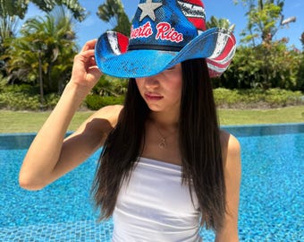 Puede incluir: Un sombrero de vaquero de paja con un diseño azul, rojo y blanco, con una estrella y el texto "Puerto Rico". El sombrero tiene un ala ancha y lo lleva una persona.