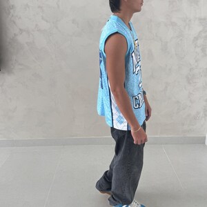 May include: A light blue basketball jersey with white and blue accents, worn with dark gray jeans and blue sneakers. The jersey has a number and letter design. The person is standing on a gray tiled floor.
