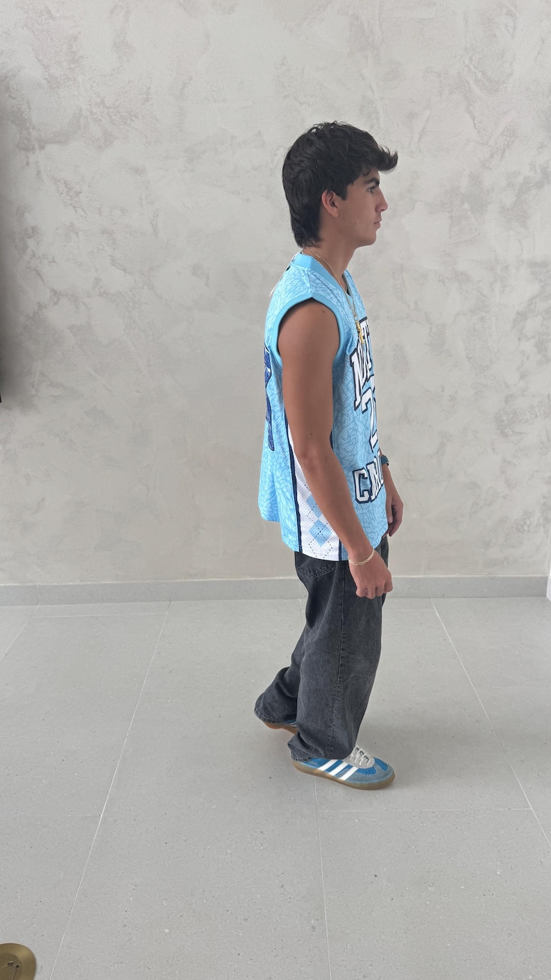 May include: A light blue basketball jersey with white and blue accents, worn with dark gray jeans and blue sneakers. The jersey has a number and letter design. The person is standing on a gray tiled floor.
