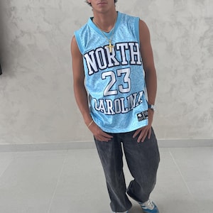 May include: A light blue basketball jersey with the text "NORTH 23 CAROLINA" is worn with dark gray pants and blue sneakers. The jersey has a mesh design. A gold necklace and watch are also visible.
