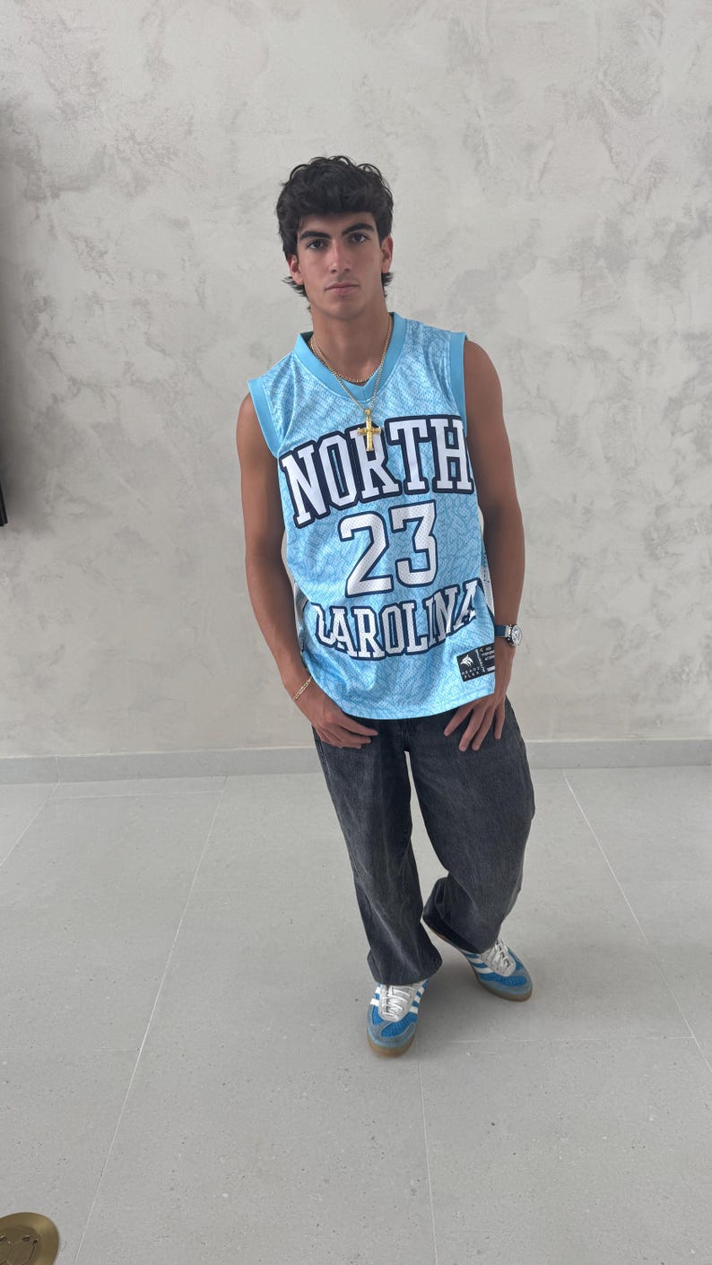 May include: A light blue basketball jersey with the text "NORTH 23 CAROLINA" is worn with dark gray pants and blue sneakers. The jersey has a mesh design. A gold necklace and watch are also visible.