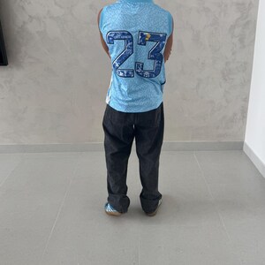May include: A person wearing a light blue basketball jersey with the number 23 in navy blue. The jersey has a patterned design and is paired with dark gray pants. The person is standing on a light gray tiled floor.