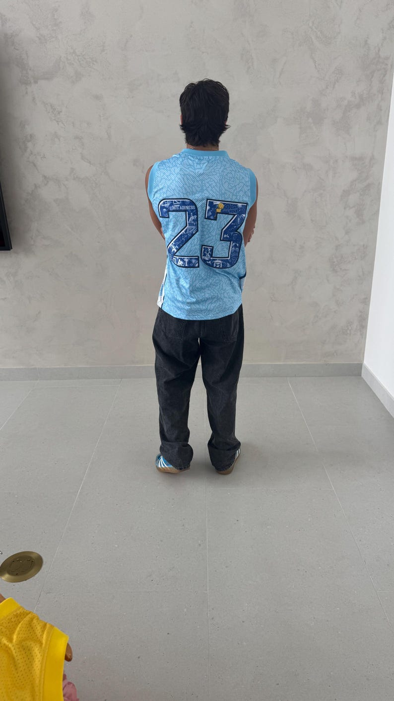 May include: A person wearing a light blue basketball jersey with the number 23 in navy blue. The jersey has a patterned design and is paired with dark gray pants. The person is standing on a light gray tiled floor.