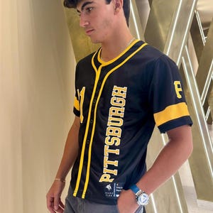 Può includere: Maglia da baseball nera e gialla con la scritta "PITTSBURGH" in verticale sul davanti. La maglia ha dettagli gialli sulle maniche e sul colletto, ed è visibile il numero "11". La maglia è indossata con pantaloni grigi e un orologio blu.