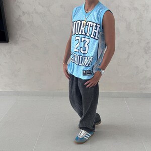 May include: A light blue basketball jersey with the text "NORTH 23 CAROLINA" is worn with dark gray jeans and blue sneakers. The jersey has a diamond pattern and a logo. A gold necklace and a watch are also visible.
