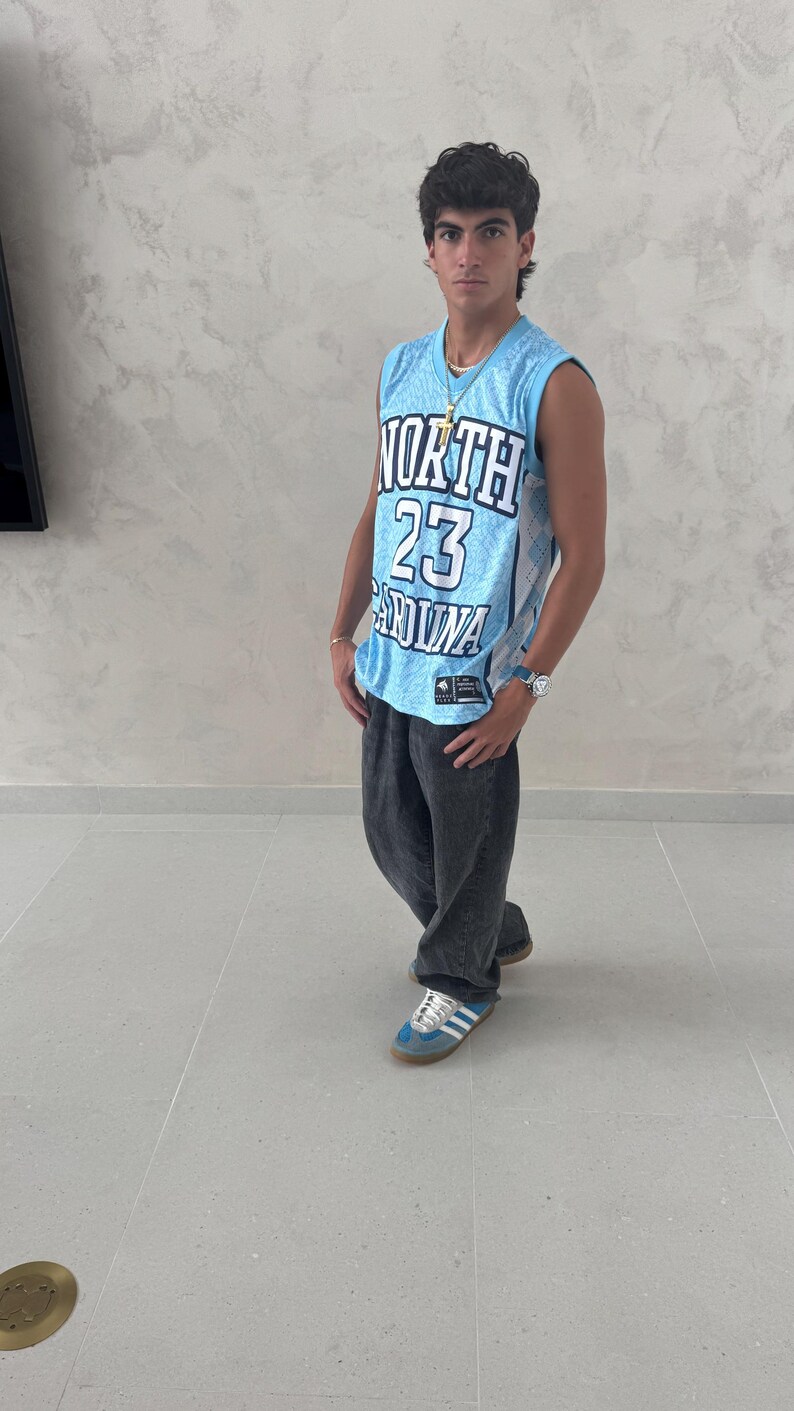 May include: A light blue basketball jersey with the text "NORTH 23 CAROLINA" is worn with dark gray jeans and blue sneakers. The jersey has a diamond pattern and a logo. A gold necklace and a watch are also visible.