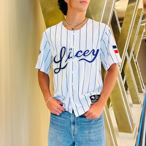 May include: A white baseball jersey with blue stripes and the word "Licey" in blue cursive lettering. The jersey has a red, white, and blue patch on the right sleeve.
