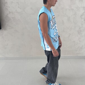 May include: A light blue basketball jersey with white and black accents, featuring the number 23 and the word "CITY". The jersey is paired with dark gray jeans and blue sneakers with white stripes.