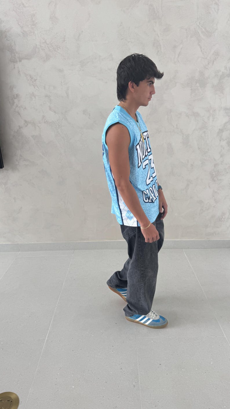 May include: A light blue basketball jersey with white and black accents, featuring the number 23 and the word "CITY". The jersey is paired with dark gray jeans and blue sneakers with white stripes.