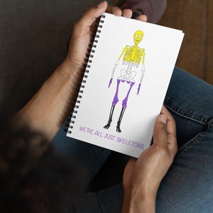Peut inclure: Un carnet à spirales blanc avec une illustration de squelette jaune et blanc portant un short violet. Le texte "We're all just skeletons" est imprimé en bas du carnet.