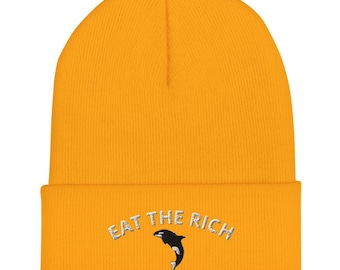 以下が含まれることがあります： 白地に「EAT THE RICH」と刺繍された黄色いビーニー。黒と白のシャチのグラフィックが描かれている。