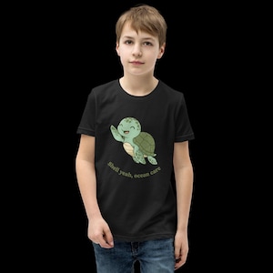 Könnte beinhalten: Schwarzes T-Shirt mit einer winkenden Zeichentrickschildkröte und dem Text "Shell yeah, ocean care." Die Schildkröte ist grün und hellbraun mit einem lächelnden Gesicht. Das T-Shirt wird von einer Person getragen.