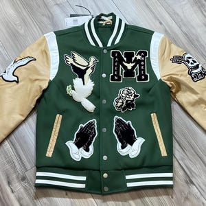 May include: Green and tan varsity jacket with multiple patches. Patches include praying hands, doves, roses, and a skull. The jacket has a white collar and cuffs with green and white stripes. The sleeves are tan.