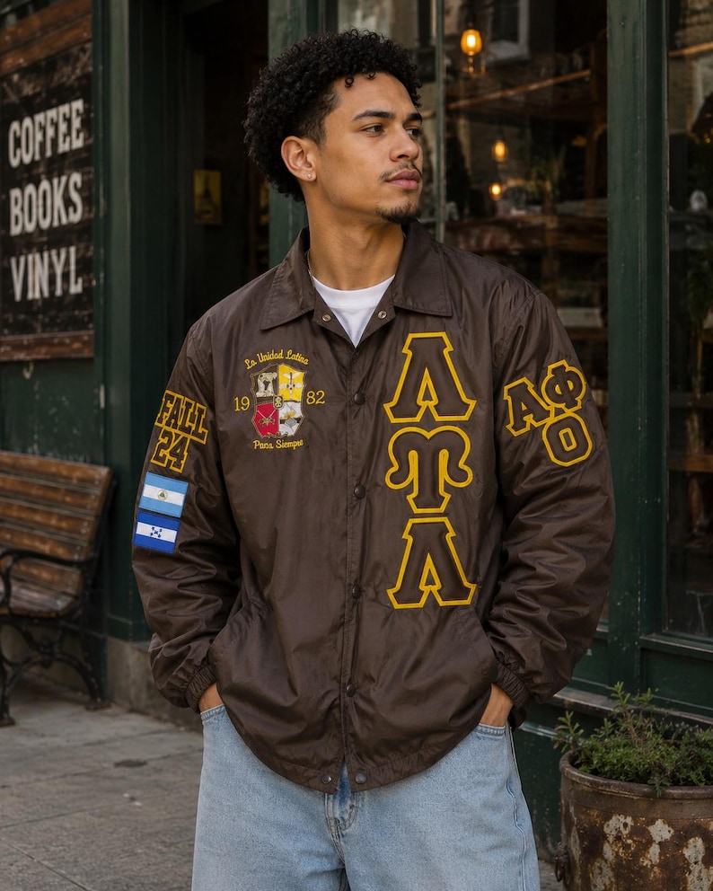 May include: A brown snap-front coach jacket featuring yellow embroidered Greek letters, the text FALL 24, and a crest with the text La Unidad Latina 1982 Para Siempre. The right sleeve displays two rectangular flags, and the left sleeve has A&Phi;&Omega;.