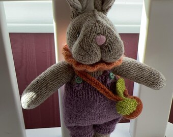 May include: A knitted gray bunny rabbit wearing purple overalls with orange straps and a green and orange leaf bag. The bunny has a pink nose and orange ruffle around its neck.