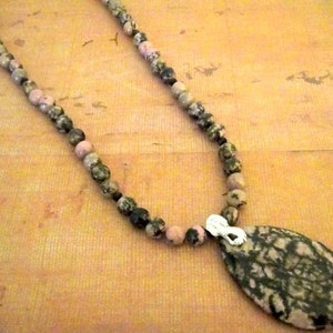May include: A pink and green stone necklace with a matching pendant. The pendant is oval shaped and has a silver clasp.