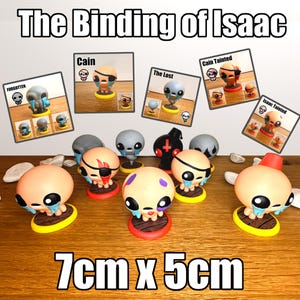 May include: A collection of figurines from "The Binding of Isaac" game, including characters like Cain and The Lost. The figurines are on wooden bases, with dimensions of 7cm x 5cm. The image also features character cards.