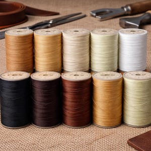 Puede incluir: Una colección de carretes de hilo en varios colores, incluyendo negro, marrón, beige y blanco. Los carretes están dispuestos en dos filas sobre una superficie de arpillera, con herramientas de marroquinería en el fondo.