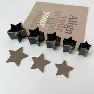 May include: Set of six metal star-shaped punch tools for crafting. The tools are arranged in order of size, with the largest star on the left and the smallest on the right.  There are also five cut-out leather stars in various sizes.
