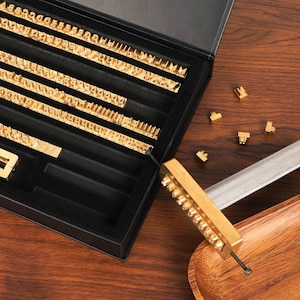 Op de afbeelding: Een vintage letterpress-drukkerskit met goudkleurige metalen letters gerangschikt in een zwarte koffer. Een metalen handvat en individuele letterblokken zijn ook zichtbaar. De kit staat op een houten oppervlak.