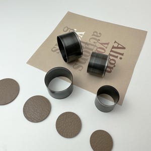 May include: Four round leather punches of different sizes, with four brown leather circles cut out by the punches. The punches are made of metal and have a silver finish. The leather circles are on a white surface. The punches are on a brown paper surface with the text "All. You. Got. This." printed on it.