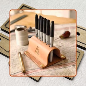 May include: A light brown leather pen holder with six black Sharpie markers. The pen holder has a curved design and is secured with brass rivets. The logo "Craftel" is visible on the front. A gold pen is in the foreground.