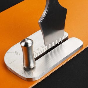 May include: A stainless steel leathercraft tool with a toothed blade and a curved handle. The tool is positioned on a piece of orange leather. The tool has measurement markings in millimetres.