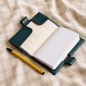 May include: Open teal leather notebook with a grid-lined notepad and a lined page. A yellow pencil with a black tip rests beside the notebook. The notebook is open on a plaid patterned surface.