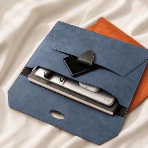 Può includere: Una custodia blu in stile busta in similpelle scamosciata con chiusura in pelle nera e fascia nera. All'interno sono presenti uno smartphone, un tablet e un taccuino. Sullo sfondo è visibile una custodia arancione.