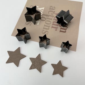 May include: Five metal star-shaped cookie cutters of varying sizes, arranged on a brown paper background.  Three brown leather star shapes are also shown.
