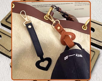 May include: Close-up of a handbag with a brown leather strap and gold-colored hardware. A black leather strap with a heart-shaped cutout and a black cap with white text are also visible. The bag has a cream-colored fabric.