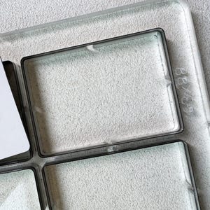 May include: A clear glass tray with four square compartments and a white card. The tray has a dark border and is set against a textured, light grey background. The dimensions are 7.6 x 7.6 cm.