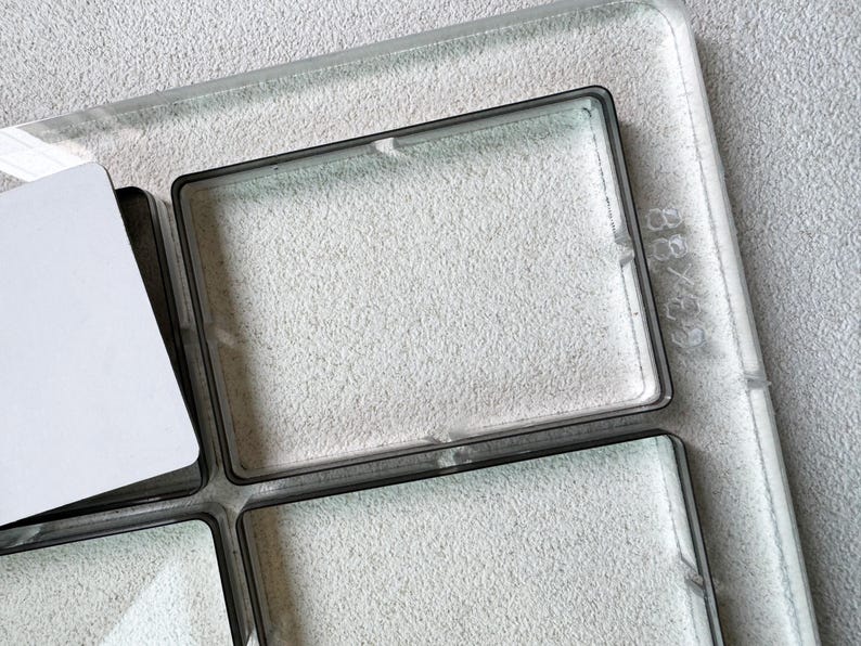 May include: A clear glass tray with four square compartments and a white card. The tray has a dark border and is set against a textured, light grey background. The dimensions are 7.6 x 7.6 cm.