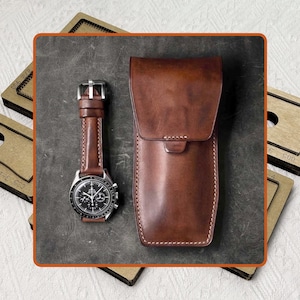 May include: A brown leather watch case and a wristwatch with a brown leather band are displayed. The watch has a black face with silver accents. The watch case is rectangular with a curved top. Wooden templates are in the background.